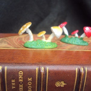 May include: A collection of five miniature mushroom figurines with white spots, red and yellow caps, and green bases. The mushrooms are arranged on a brown leather-bound book with gold accents and the text "The Week-End Book".