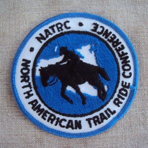 Può includere: Una toppa ricamata blu e bianca con il testo "North American Trail Ride Conference" e "NATRC" attorno a un cerchio blu con una silhouette bianca di un cavaliere a cavallo.