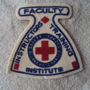 Puede incluir: Un parche de fieltro blanco con bordados azules y rojos. El parche tiene forma de triángulo con la parte superior redondeada. Dice "FACULTY INSTRUCTORS SAFETY SERVICES TRAINING INSTITUTE" con una cruz roja en el centro.