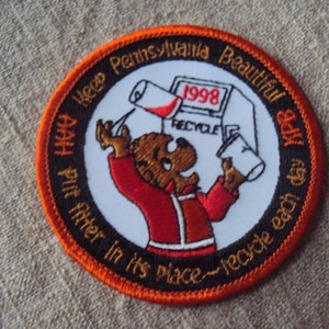 Pode incluir: Remendo circular com borda laranja e o texto "Keep Pennsylvania Beautiful". O remendo apresenta um urso de desenho animado com uma jaqueta vermelha reciclando. O ano de "1998" é exibido. O texto "AAH Put filter in its place~recycle each day" também está incluído.