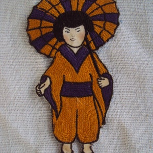 Peut inclure: Patch brodé représentant une personne portant un kimono jaune et violet avec un parapluie violet et orange. La personne a les cheveux noirs et une expression neutre.