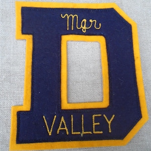 Könnte beinhalten: Ein marineblaues Filzbuchstabe "D" mit einem goldenen Rand. Das Wort "Mgr" ist oben und "VALLEY" unten mit goldfarbenem Faden gestickt. Der Aufnäher ist wahrscheinlich für eine Collegejacke.