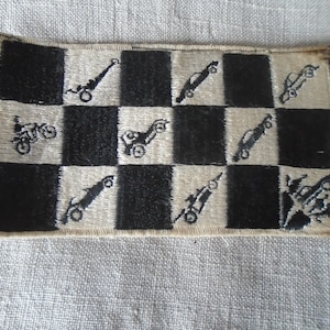 May include: A rectangular patch with a checkered pattern of black and beige squares. The beige squares feature embroidered black images of vehicles, including motorcycles, cars, and what appears to be a cannon. The patch has a beige border.