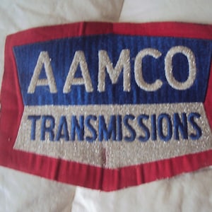 May include: A rectangular patch with a red border, blue center, and silver and white text. The text reads "AAMCO TRANSMISSIONS". The patch is likely for a uniform or promotional item.