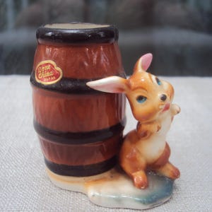 Może przedstawiać: Ceramiczna figurka brązowej beczki z czarnymi pasami i królikiem. Królik ma pomarańczowo-białe futro, różowe uszy i niebieskie oczy. Beczka ma naklejkę "Bone China Japan".