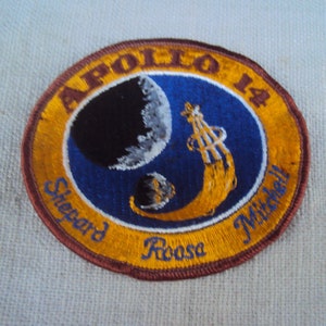 May include: Circular Apollo 14 patch with a blue background, featuring a stylized rocket and celestial bodies. The patch has a gold border and text that reads "Apollo 14", "Shepard", "Roosa", and "Mitchell".