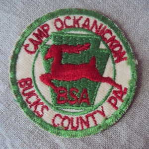 May include: A green and white embroidered patch with the text "Camp Ockanickon Bucks County BSA PA". A red deer is leaping in the center of the patch.
