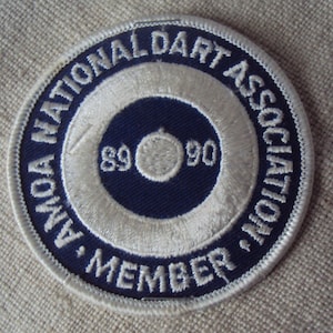 以下が含まれることがあります： 縁に「AMOA NATIONAL DART ASSOCIATION MEMBER」という文字が刺繍された白と青のワッペン。ワッペンの中央には、白い的と「89」と「90」の数字が書かれた青い円があります。