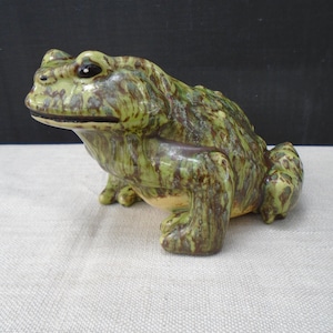 May include: A ceramic frog figurine featuring a glossy glaze with mottled shades of olive green and dark brown. The sculpture depicts a frog in a seated position with textured skin, prominent eyes, and a wide mouth, showcasing a detailed finish.