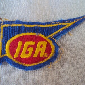 May include: A blue and red embroidered patch with the word "IGA" in yellow lettering.