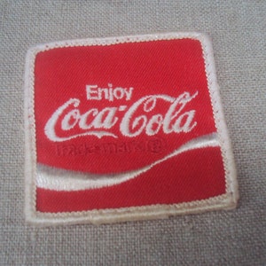 Può includere: Toppa ricamata rossa con cuciture bianche. La toppa presenta il logo Coca-Cola e il testo "Enjoy Coca-Cola Trademark Reg."