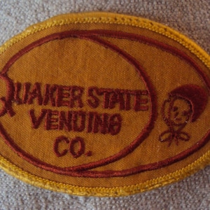 Könnte beinhalten: Ein brauner und gelber ovaler Patch mit dem Text "Quaker State Vending Co." in roter Stickerei. Eine kleine rote Comicfigur befindet sich auf der rechten Seite des Patches.