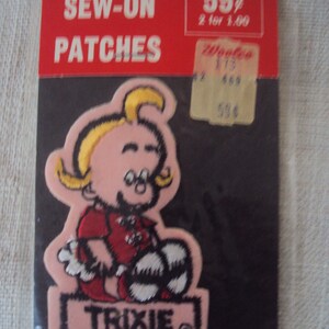 Może przedstawiać: Naszywka do przyszycia w kolorze różowym i białym przedstawiająca kreskówkową dziewczynkę z blond włosami, ubraną w czerwoną koszulę i trzymającą piłkę nożną. Naszywka zawiera napis "TRIXIE" na różowym banerze.