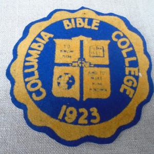 Puede incluir: Un parche vintage del Columbia Bible College con un centro azul y un borde festoneado amarillo. El parche presenta las palabras "Columbia Bible College" y "Bible College" en amarillo, junto con el año "1923". El centro tiene una cruz y otros gráficos.
