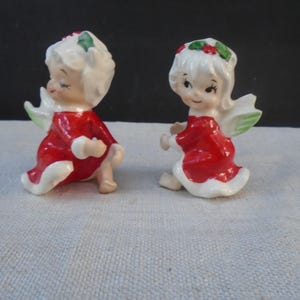 Può includere: Due statuette di angeli in ceramica con capelli bianchi decorati con agrifoglio verde e bacche rosse. Ogni angelo indossa un abito rosso brillante con bordo bianco e ha piccole ali verde pallido. Le figure sono raffigurate in posizione seduta.