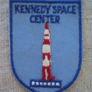 Könnte beinhalten: Ein blauer und weißer gestickter Aufnäher mit dem Text "Kennedy Space Center" und einer roten und weißen Rakete.