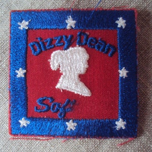 Puede incluir: Un parche bordado rojo y azul con estrellas blancas alrededor del borde. El centro presenta una silueta blanca de una niña con el texto "Dizzy Dean" arriba y "So" abajo.