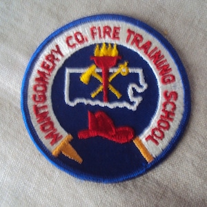 May include: A blue and white embroidered patch with the text "Montgomery Co. Fire Training School" in a circle. The patch features a red fire hat, crossed axes, and a torch.