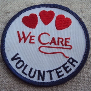 Può includere: Una toppa ricamata bianca con un bordo blu. La toppa ha tre cuori rossi e il testo "We Care Volunteer".
