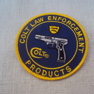 May include: A round patch with a navy blue background and yellow border. The patch features the text "COLT LAW ENFORCEMENT PRODUCTS" in yellow. A silver and black handgun is in the center, with the Colt logo in yellow below it.