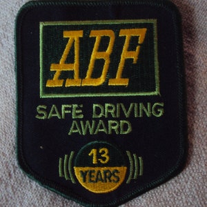 Könnte beinhalten: Ein gestickter schwarzer und grüner Aufnäher mit dem Text "ABF Safe Driving Award" und einem gelben Kreis mit dem Text "13 Years".