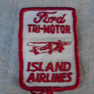 May include: A rectangular patch with a white background and red trim. The patch features the words "Ford TRI-MOTOR" and "ISLAND AIRLINES" in red embroidered lettering, along with a red embroidered airplane design.