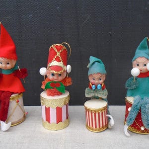 Puede incluir: Cuatro adornos navideños vintage de elfos. Cada elfo está sentado en un tambor y viste un atuendo de diferente color. Los elfos sostienen pompones blancos. Los tambores son rojos, blancos y dorados.