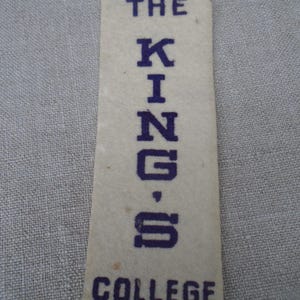 Könnte beinhalten: Ein rechteckiges, cremefarbenes Etikett mit den Worten "THE KINGS COLLEGE" in Dunkelblau gedruckt. Der Text ist vertikal ausgerichtet und auf dem Etikett zentriert. Das Etikett scheint aus Papier oder einem ähnlichen Material zu bestehen.