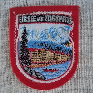 May include: A red felt patch with white stitching around the edges. The patch features a white background with a silver embroidered image of a mountain range and a building. The text "EIBSEE MIT ZUGSPITZE" is embroidered in silver on the top of the patch.