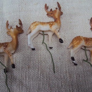 May include: Three plastic deer figurines with brown and white paint, each with a green wire stem attached to the bottom.