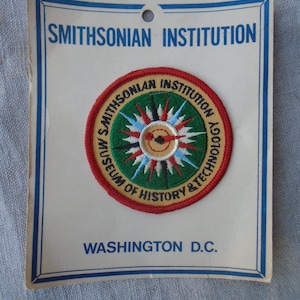 May include: A white ceramic mug with a black handle and the text "But First, Coffee" in a black, bold font. The mug is on a white surface. The mug is approximately 10 cm tall.