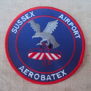 May include: A blue and red embroidered patch with the text "SUSSEX AIRPORT AEROBATEX" and a gray eagle with a red, white, and blue airplane underneath.