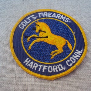 May include: Embroidered patch featuring a yellow horse leaping within a blue circle, encircled by text reading "COLTS FIREARMS HARTFORD, CONN" in white. The patch has a yellow border and is likely a vintage collectible.