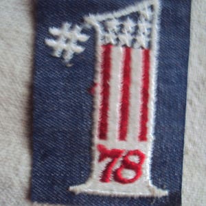 Puede incluir: Un parche de mezclilla azul con un número uno blanco bordado en él. El número uno está decorado con rayas rojas y blancas que se asemejan a una bandera estadounidense. El número 78 está bordado en rojo debajo del número uno.