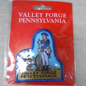 以下が含まれることがあります： 白い文字でVALLEY FORGE PENNSYLVANIAと書かれた赤い長方形のカード。青い縁取りの刺繍ワッペンには、青いコートを着て大砲のそばに立つ兵士が描かれており、下部にはVALLEY FORGE PENNSYLVANIAの文字が記されている。