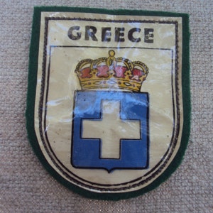 Könnte beinhalten: Ein grüner Filz-Patch mit weißem Hintergrund und dem Wort "GREECE" in schwarzen Buchstaben. Der Patch zeigt ein blaues Schild mit einem weißen Kreuz und einer goldenen Krone über dem Schild.