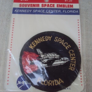 以下が含まれることがあります： フロリダ州ケネディ宇宙センターの記念宇宙エンブレム。黒い円形のパッチには、スペースシャトルのデザインと「Kennedy Space Center Florida」と書かれた金色の文字が特徴です。パッチは、赤と白のヘッダーが付いた透明なプラスチック製のスリーブにパッケージされています。