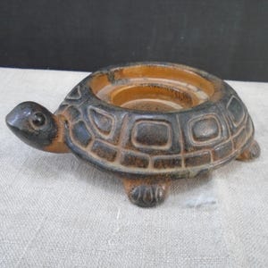 May include: A ceramic turtle-shaped candle holder. The candle holder is brown and black with a textured shell design. The turtle has a small head and four stubby legs. The candle holder is designed to hold a single candle.