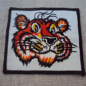 Może przedstawiać: Haftowana naszywka przedstawiająca stylizowaną twarz tygrysa. Twarz tygrysa jest głównie pomarańczowa, czerwona i żółta, z czarno-białymi akcentami. Naszywka jest kwadratowa z czarną obwódką i na białym tle.
