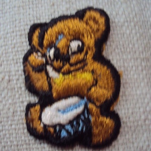 May include: Embroidered patch of a brown bear playing a drum. The bear is wearing a yellow hat and has a blue and white drum.