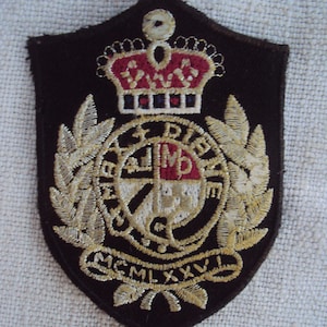 Könnte beinhalten: Ein schwarzer gestickter Aufnäher mit einer goldenen Krone, einem Schild und einem Lorbeerkranz. Der Schild hat ein rotes und weißes Design mit den Buchstaben "D.M.D." und dem Jahr "MCM LXXV" in Gold.