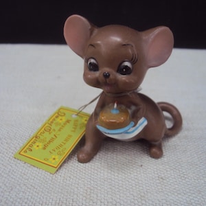 May include: A brown ceramic mouse figurine holding a small birthday cake with a blue and white stripe. The figurine has large ears and a tag that reads "Happy Birthday" and "Morse Village".