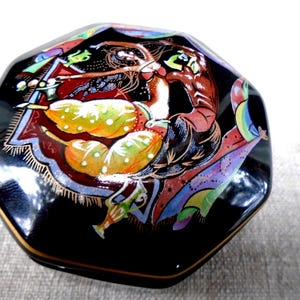 May include: A black, octagonal trinket box with a gold rim. The lid features a colorful illustration of a figure with flowing hair and a patterned design. The artwork includes shades of red, yellow, and blue.