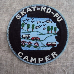 May include: A blue and black embroidered patch with the text "SKAT-RD-FU CAMPER" and a cartoon image of a landscape with trees, clouds, and vehicles.
