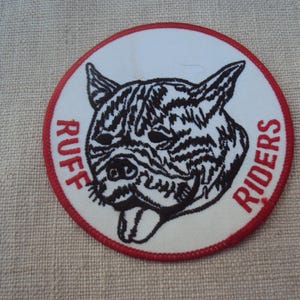 May include: Circular patch with a white center featuring a black and white dog face design. The words "RUFF RIDERS" are in red around the edge of the patch, which has a red border.
