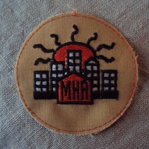 May include: A tan circular patch with an embroidered design of a red building with black windows and a black sun with rays behind it. The building has the letters "MHA" in red on the front.