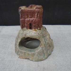 May include: A small, textured ceramic ornament shaped like a castle tower perched atop a rocky base. The tower is a deep reddish-brown color, while the base is a mottled, pale greenish-gray with a large, hollow opening carved into the front side.