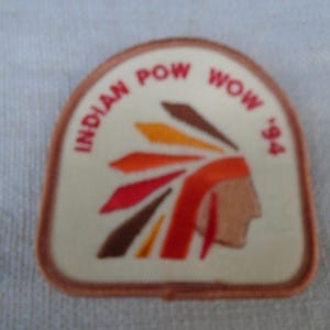 Op de afbeelding: Een crèmekleurige geborduurde patch met een bruine rand. De patch toont het profiel van een persoon met een hoofdtooi met oranje, gele, rode en bruine veren. De tekst "INDIAN POW WOW '94" is in het rood langs de bovenrand gedrukt.