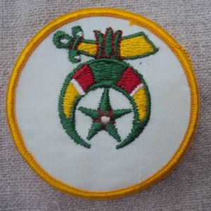 May include: A white embroidered patch with a yellow border. The patch features a green crescent moon with a five-pointed star in the center. The crescent moon is topped with a red and yellow design.