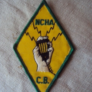 Pode incluir: Um remendo amarelo em forma de diamante com uma borda verde. O remendo apresenta uma mão segurando um microfone com o texto "NCHA C.B." em preto.